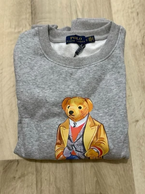 Grå sweatshirt Polo Bear Ralph Lauren - Grå sweatshirt från Polo Ralph Lauren med ikonisk Polo Bear-tryck på bröstet. Björnen har en gul kavaj, röd tröja och blå jeans, samt en shoppingpåse. Tröjan har rund hals, ribbade muddar och mjukt material som känns skönt mot huden.