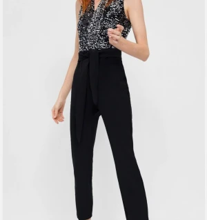 Svart jumpsuit med paljetter och knyt - Snygg svart jumpsuit med croppade ben och högt knyt i midjan. Överdelen är täckt av glittriga silverpaljetter och har en djup v-ringning. 