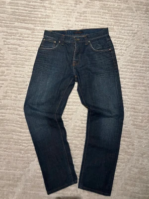 Mörkblå jeans från Nudie Jeans - Snygga mörkblå jeans från Nudie Jeans med klassisk femficksdesign och orange kontrastsömmar. Baktill finns Nudie Jeans typiska broderade vågmönster på fickan och en läderpatch i midjan. Jeansen har ”steady Eddie” passform och är tillverkade i slitstarkt denimtyg.