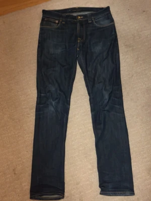 Mörkblå Nudie Jeans Co jeans W31 L32 - Snygga mörkblå jeans från Nudie Jeans Co i ekologisk denim. Modellen har klassisk femficksdesign, kontrastsömmar och Nudie-logga på bakfickan. Jeansen är raka i benen och har normal passform. Perfekt för dig som gillar stilrena och hållbara jeans.