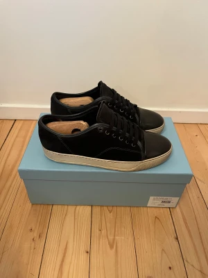 Svarta sneakers från Lanvin i mocka - Lanvin sneakers i svart mocka med rund tå och platt vit sula. Skorna har snörning och detaljer i svart skinn på tån. Klassisk och stilren design med metallöglor och diskret söm längs sidan. Perfekt för dig som gillar lyxiga och minimalistiska sneakers.