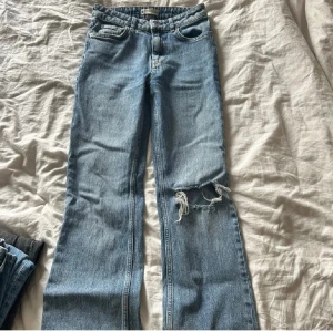 Blå jeans med hål på knät - Säljer ett par klassiska blå jeans med rak passform och snyggt slitet hål på ena knät. Jeansen har fem fickor, knappgylf och är i ett mjukt denimtyg. Perfekt för dig som gillar en avslappnad och cool stil.