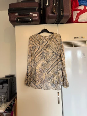 Mönstrad långärmad skjorta från odd Molly  - Säljer en unik långärmad skjorta från Odd Molly med ett detaljerat paisley- och randigt mönster i beige, grått och svart. Skjortan har v-ringad hals, knappar framtill och är tillverkad i ett mjukt, tunt material som känns skönt mot huden. Från märket Odd Molly. Storlek 3 som enligt deras storleksguide motsvarar en L. Mycket fint skick, jag hittar inga synliga anmärkningar eller defekter. 