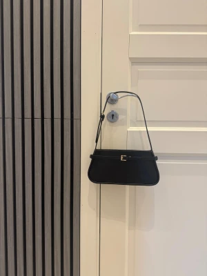 Svart axelväska med gulddetalj - Snygg svart axelväska i stilren design med rektangulär form och smal axelrem. Väskan har en guldfärgad spänndetalj framtill och dragkedja upptill. 