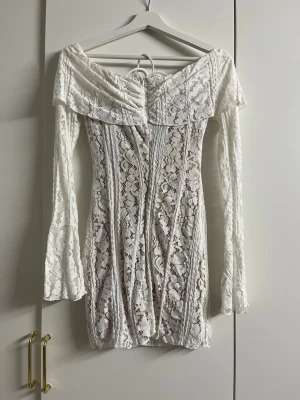 Vit spetsklänning med snörning - Superfin vit kortklänning i spets med offshoulder och långa trumpetärmar. Klänningen har snörning framtill och ett romantiskt blommigt spetsmönster. Perfekt för sommarens alla tillfällen och ger en boho-vibe.