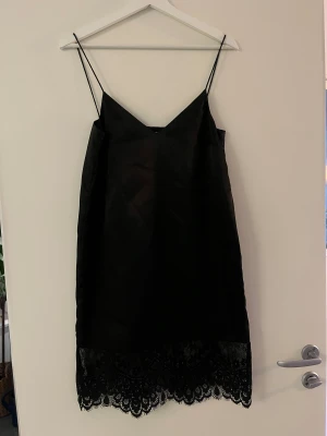 Svart spetsklänning från Mango XS - Svart kortklänning från Mango med tunna axelband och v-ringad hals. Klänningen har en slät satinliknande yta och avslutas med bred spetskant nedtill som ger en extra snygg touch. Perfekt för dig som gillar stilrena och eleganta plagg.