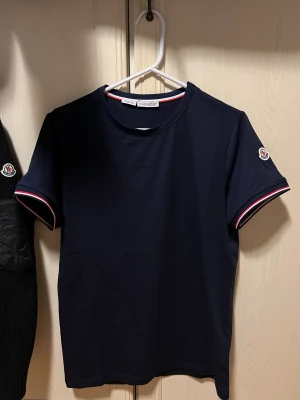 Mörkblå Moncler t-shirt slim fit - Snygg mörkblå t-shirt från Moncler i slim fit-modell. Klassisk design med korta ärmar och Moncler-logga på ärmen. Ärmsluten har stilrena ränder i vitt och rött. Tillverkad i mjuk bomull som känns skön mot huden. Storlek S, felfri och aldrig använd.