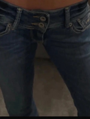 Blå low waist jeans från Pepe Jeans - Säljer ett par blå jeans från Pepe Jeans med låg midja och raka ben. Jeansen har klassisk femficksdesign, dubbla knappar i midjan och snygga slitningar för en avslappnad look. Jeansen är ganska stretchiga så de passar s/m skulle jag säga!