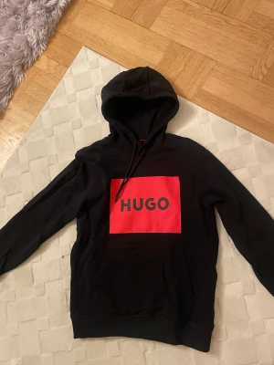 Svart hoodie från Hugo med röd logo - Svart hoodie från Hugo med stor röd logga på bröstet och svarta snören i huvan. Klassisk passform med känguruficka framtill. Mjuk bomullskänsla och stilren design som sticker ut med den feta röda rutan och tydlig HUGO-text. Använt den ett fåtal gånger och har annars bara legat hemma.