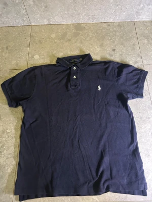 Mörkblå Polo Ralph Lauren t-shirt - Snygg mörkblå pikétröja från Polo Ralph Lauren i custom slim fit. Klassisk pikémodell med krage, två vita knappar och broderad logga på bröstet. Skulle säga att den är lite urtvättat men inget man tänker på!