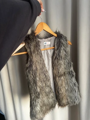Grå pälsväst från H&M - Säljer en fluffig och mjuk grå pälsväst från H&M. Västen har en öppen front och är fodrad med slätt tyg på insidan. Pälsen är syntetisk och har en naturlig grå ton med mörkare inslag för en cool look.