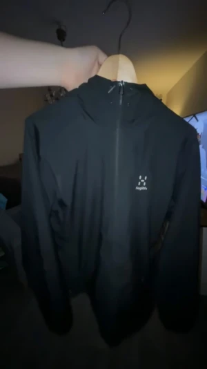 Svart vindjacka från Haglöfs - Svart vindjacka från Haglöfs med minimalistisk design och diskret logga på bröstet. Jackan har huva och hel dragkedja framtill, perfekt för blåsiga dagar. Lätt och smidig att bära, passar dig som gillar outdoor och streetstyle.