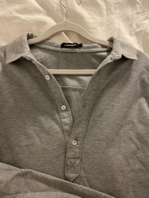 Grå henley-skjorta från J.Lindeberg - Snygg grå henley-skjorta från J.Lindeberg med klassisk krage och tre knappar framtill. Skjortan är långärmad och tillverkad i mjuk bomull. Passar på en vardag.