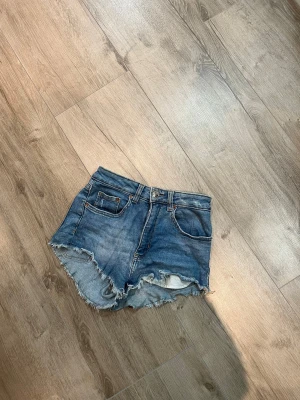 Blå jeansshorts från H&M Divided - Säljer ett par blå jeansshorts från H&M Divided i storlek 36. De har råa, fransiga kanter och slitna detaljer framtill för en cool, avslappnad look. Nyskick.