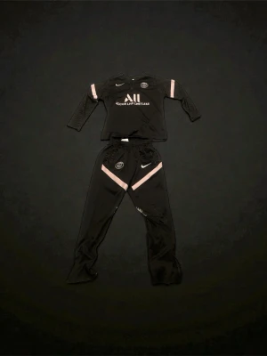 PSG x Nike svart träningsset - Svart träningsset från Nike och PSG med långärmad tröja och byxor. Setet har rosa detaljer, PSG-logga och Nike Swoosh på både tröja och byxor. Materialet är lätt och syntetiskt, perfekt för fotbollsträning. Tröjan har half zip och byxorna har resår i midjan. Hela setet går för 600 kronor. Men du kan såklart också betala för de separat bara för Tröjan kostar det 400 kr och för byxorna separat 200kr. Storlek är 158-164 som då passar 13-14 år