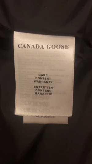 Svart dunjacka från Canada Goose M - Säljer en svart dunjacka från Canada Goose i storlek M. Jackan har klassisk pufferstil med huva och det ikoniska röda Canada Goose-märket på ärmen. Perfekt för kalla dagar och riktigt varm. Stängs med dragkedja och tryckknappar plus QR-Kod och NFC-tac