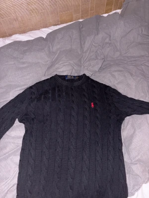 Svart stickad tröja Polo Ralph Lauren - Svart kabelstickad tröja från Polo Ralph Lauren med klassisk rund hals och röd broderad logga på bröstet. Tröjan har långärmad passform och ribbade muddar vid hals, ärmslut och nederkant. Perfekt för dig som gillar stilren och tidlös design.