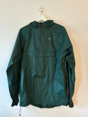 Grön vindjacka från Packmack M - Mörkgrön vindjacka från Packmack i storlek M. Jackan har huva, half zip-dragkedja framtill och en stor ficka med dragkedja på magen. Tillverkad i 100% nylon, perfekt för blåsiga dagar. Snygg minimalistisk design med logga på bröstet.