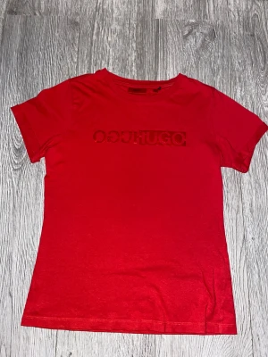 Röd HUGO t-shirt med logga - Snygg röd t-shirt från HUGO med diskret logotyp i rött på bröstet. Modellen är klassisk med rund hals och korta ärmar. Tillverkad i 100% ekologisk bomull som känns mjuk mot huden. Perfekt för dig som gillar stilrena och trendiga plagg.