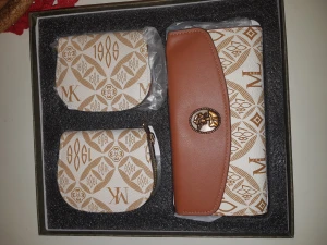 Set med plånbok och småväskor MK - Snyggt set med en plånbok och två småväskor från MK. Designen är vit och beige med bruna detaljer och mönster med logotyp och siffror. Plånboken har ett brunt lock med guldfärgad knapp och stilren form. Perfekt för att hålla ordning på kort och småsaker.
