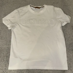 Vit Burberry t-shirt med logga - Vit t-shirt från Burberry med klassisk rund hals och upphöjd Burberry-logga framtill. Tillverkad i mjuk bomull och har korta ärmar. Snygg och stilren design med diskreta detaljer i halsringningen.