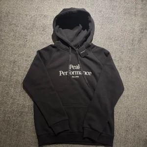 Svart hoodie från Peak Performance - Svart hoodie från Peak Performance med vit broderad logga och text 'Est. 1986' på bröstet. Klassisk modell med huva och snörning, samt mjukt material och ribbade muddar. Perfekt för en chill och stilren look.