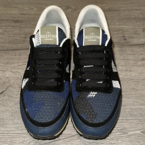 Valentino Garavani Rockrunner sneakers blå - Snygga Valentino Garavani Rockrunner sneakers med blått och svart camouflage-mönster. Skorna har svarta snören, beige sula och vita detaljer vid hälen. Ovandelen är i textil och mocka, med en robust gummisula för extra grepp. Perfekt för dig som gillar exklusiva streetwear-vibes.