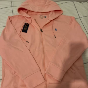 Ljusrosa hoodie från Polo Ralph Lauren - Snygg ljusrosa hoodie från Polo Ralph Lauren med dragkedja och huva. Klassisk blå logga broderad på bröstet och praktiska fickor framtill. Tillverkad i mjuk bomull, perfekt för chill dagar. Stilsäker och enkel att matcha med jeans eller joggers.