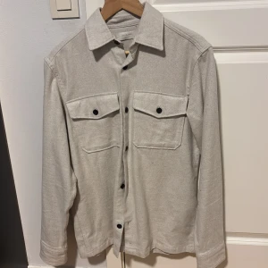 Beige overshirt från Jack & Jones - Stilren beige overshirt från Jack & Jones Premium med svarta knappar och två stora bröstfickor. Skjortan har klassisk krage och är tillverkad i ett mjukt bomullstyg. Perfekt lager-på-lager-plagg med relaxed vibe.