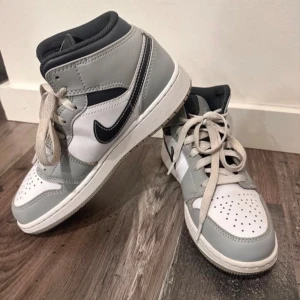 Nike Air Jordan 1 Mid Grå/Vit - Säljer ett par Nike Air Jordan 1 Mid i grått och vitt med svarta detaljer. Klassisk high-top siluett med snörning och perforerad tå. Skorna har en vit bas med ljusgrå overlays och svart swoosh. Perfekta för dig som gillar streetstyle och vill sticka ut. Pris kan diskuteras.
