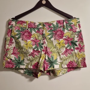 Blommiga shorts från H&M, strl 40 - Färgglada shorts från H&M med tropiskt blommönster i rosa, gul och grönt på vit botten. Klassisk passform med bälteshällor och fickor, perfekta för sommardagar. Lätta och sköna, tillverkade i bomull.