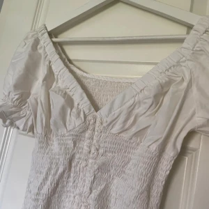 Vit smockad kortklänning med puffärm - Supersöt vit kortklänning med smockad kropp och puffiga korta ärmar. Klänningen har v-ringning och volangdetaljer vid ärmslut. 