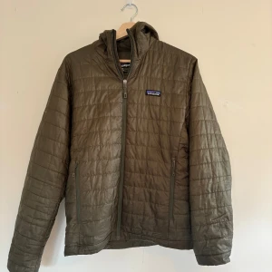 Olivgrön funktions jacka från Patagonia - Säljer en olivgrön  pufferjacka från Patagonia i storlek S. Jackan har huva, hel dragkedja framtill och två sidofickor med dragkedja. Tillverkad i lätt polyester och perfekt för kyliga dagar. Klassisk logga på bröstet och stilren look.