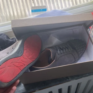 Christian Louboutin gråa sneakers med nitar - Säljer ett par gråa sneakers från Christian Louboutin med ikonisk röd sula och coola nitar på tån. Skorna har snörning och är tillverkade i mocka med läderdetaljer. Kommer med originalkartong och kvitto. Perfekta för dig som vill sticka ut med lyxig streetstyle.