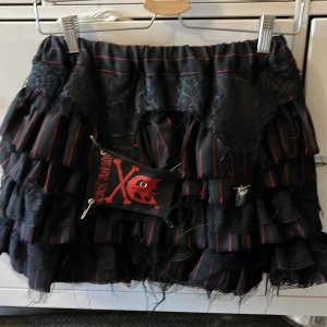 Svart volangkjol från Hell Bunny - Svart kortkjol från Hell Bunny med röda ränder, flera lager volanger och trasiga spetsdetaljer. Kjolen har säkerhetsnålar och en cool patch framtill med döskallekanin. Perfekt för dig som gillar alternativ stil och vill sticka ut.