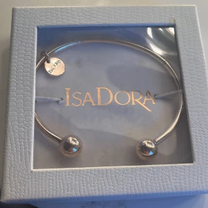 Guldfärgat armband från IsaDora - Stilrent guldfärgat armband från IsaDora med minimalistisk design. Armbandet är öppet och pryds av två blanka kulor i ändarna samt en liten rund bricka med IsaDora-loggan. Perfekt accessoar för en clean look.