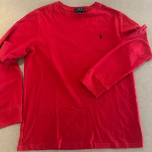 Röd långärmad tröja från Polo Ralph Lauren - Klassisk röd långärmad tröja från Polo Ralph Lauren med rund halsringning och ikonisk blå broderad logga på bröstet. Tillverkad i mjuk bomull som är skön mot huden. Perfekt för dig som gillar stilrena och tidlösa plagg.