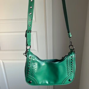 Grön axelväska med kedjedetalj - Cool grön axelväska i syntetmaterial med silverfärgade nitar och kedja. Väskan har justerbart axelband med metallringar och dragkedja upptill. Snygga sömdetaljer på framsidan ger extra edge. Perfekt storlek för det viktigaste och en riktigt trendig look.