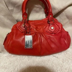 Röd handväska från Marc by Marc Jacobs - Säljer en snygg röd handväska från Marc by Marc Jacobs i äkta skinn. Väskan har silverfärgade metalldetaljer, dubbla handtag och avtagbar axelrem. Insidan är fodrad med svartvitt mönster och har dragkedja. Perfekt för dig som vill sticka ut med en färgstark accessoar.