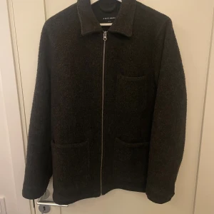 Mörkgrå overshirt från A Day's March - Säljer en stilren mörkgrå overshirt från A Day's March i ullmix. Jackan har rak passform, tre fickor framtill och stängs med dragkedja. Perfekt för lager-på-lager och funkar året runt. Enkel och clean design som passar till allt.