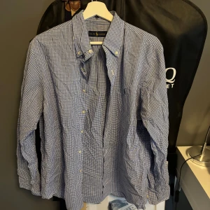 Blårutig skjorta från Ralph Lauren L - Klassisk blårutig skjorta från Ralph Lauren i storlek L. Skjortan har button down-krage, långa ärmar och vita knappar framtill. Tillverkad i mjuk bomull och har en snygg passform som funkar till många olika stilar.