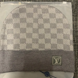 Gråvit rutig Louis Vuitton mössa - Exklusiv mössa från Louis Vuitton i grått och vitt med rutigt mönster och bred uppvikt kant. LV-logga framtill. Stickad i mjukt material som känns lyxigt och passar perfekt när du vill ha en clean och stilren look.