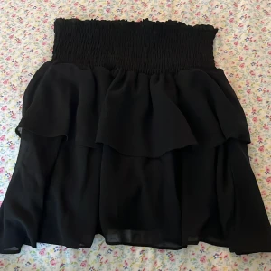 Svart volangkjol från Chelsea - Supersöt svart kortkjol från Chelsea med bred smock i midjan och dubbla volanger. 💗💗Den går även att använda som tub topp💗Säljer för att jag inte använder den! Priset kan diskuteras! 