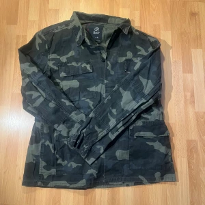 Camouflage overshirt från Brandit XL - Säljer en grön och svart camouflage overshirt från Brandit i storlek XL. Jackan har klassisk krage, flera fickor framtill och knäppning med knappar. Perfekt för dig som gillar streetstyle och vill sticka ut med en militärinspirerad look.