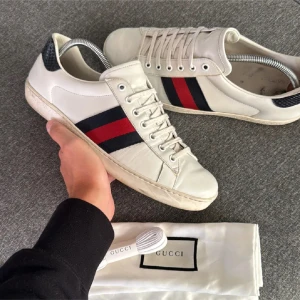 Gucci vita sneakers med röd/blå rand - Gucci sneakers i vitt skinn med klassiska röd och marinblå ränder på sidorna. Baksidan har svart ormskinnsmönstrad detalj med Gucci-logga. Platt sula, snörning och rund tå. Snygga och ikoniska skor som lyfter varje outfit. Pris kan diskuteras.🙌