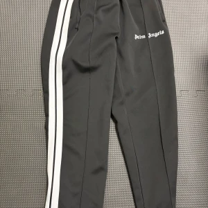 Palm Angels svarta trackpants med ränder - Svarta trackpants från Palm Angels med vita dubbla sidoremsor och logga tryckt på låret. Byxorna har elastisk midja och är tillverkade i ett glansigt syntetmaterial som ger en sportig vibe. Perfekta för en streetwear-look. Vill du ha mer bilder skriv i dm