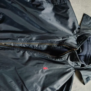 Blå vindjacka från Polo Ralph Lauren - Svart vindjacka från Polo Ralph Lauren med klassisk röd logga broderad på bröstet. Jackan har dragkedja i guldton, två fickor framtill och mjukt fleecefoder på insidan. Perfekt för dig som vill ha en stilren och tidlös look. Stor i storlek