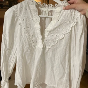 Vit blus med broderad spetskrage H&M - Superfin vit blus från H&M med stor broderad spetskrage och dekorativa knappar framtill. Långärmad modell i mjuk bomull med romantiska detaljer och v-ringning. Perfekt för dig som gillar vintagevibbar och söta detaljer.