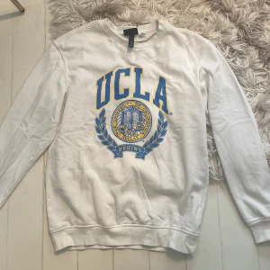 Vit UCLA sweatshirt med tryck - Säljer en vit sweatshirt från H&M med stort blått och gult UCLA-tryck på bröstet. Tröjan har rund halsringning och långa ärmar. Perfekt för dig som gillar college-stil och vill ha en skön och snygg tröja till vardags.