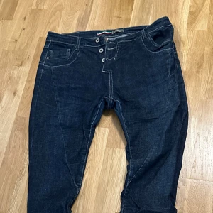 Mörkblå jeans från Please Vintage Chic - Snygga mörkblå jeans från Please Vintage Chic med vita kontrastsömmar, knäppning med tre knappar och klassiska fem fickor. Bakfickorna har en unik design med ett litet hjärtdetalj på ena fickan. Perfekta för dig som gillar italiensk streetstyle.